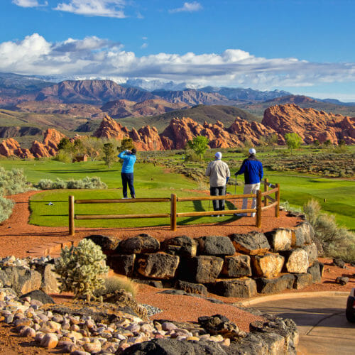 Group of three men teeing off on desert golf course with red rock formation in distance and white clouds in a bright blue sky.
